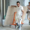 Mondelin 900kg Board Trolley – Tradesmen Moving Plasterboard Two tradesmen transporting heavy plasterboard sheets across a site using the Mondelin 900kg board trolley available for hire from Weller Hire.