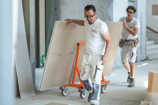 Mondelin 900kg Board Trolley – Tradesmen Moving Plasterboard Two tradesmen transporting heavy plasterboard sheets across a site using the Mondelin 900kg board trolley available for hire from Weller Hire.